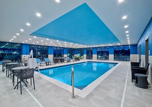 An indoor swimming pool with a blue ceiling, surrounded by lounge chairs and tables, and large windows offering city views. - Hampton Inn by Hilton Omaha UN Medical Center Area (Omaha)