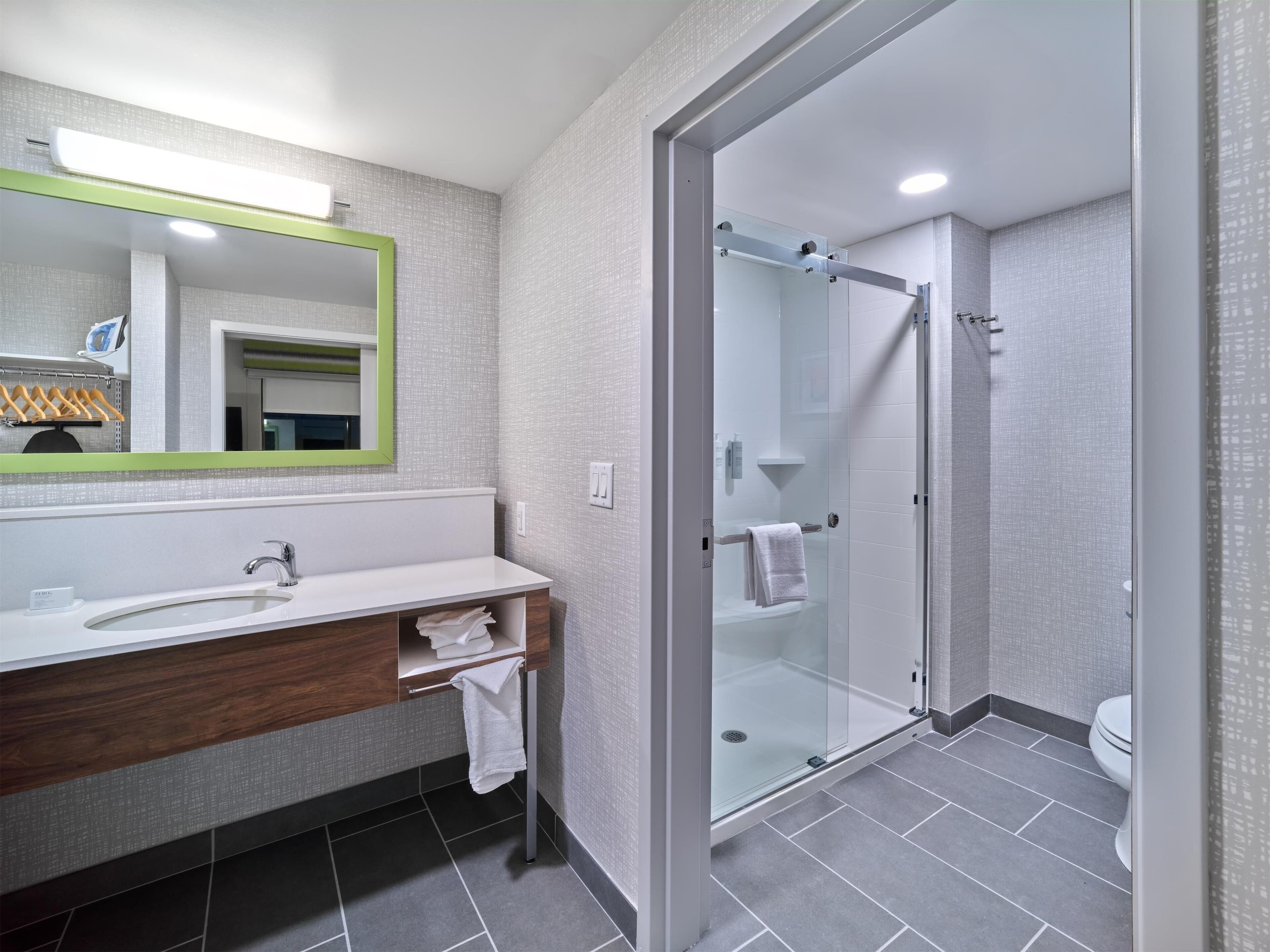 A modern bathroom with a glass shower enclosure, a large mirror with a green frame, and a white sink with a wooden vanity.