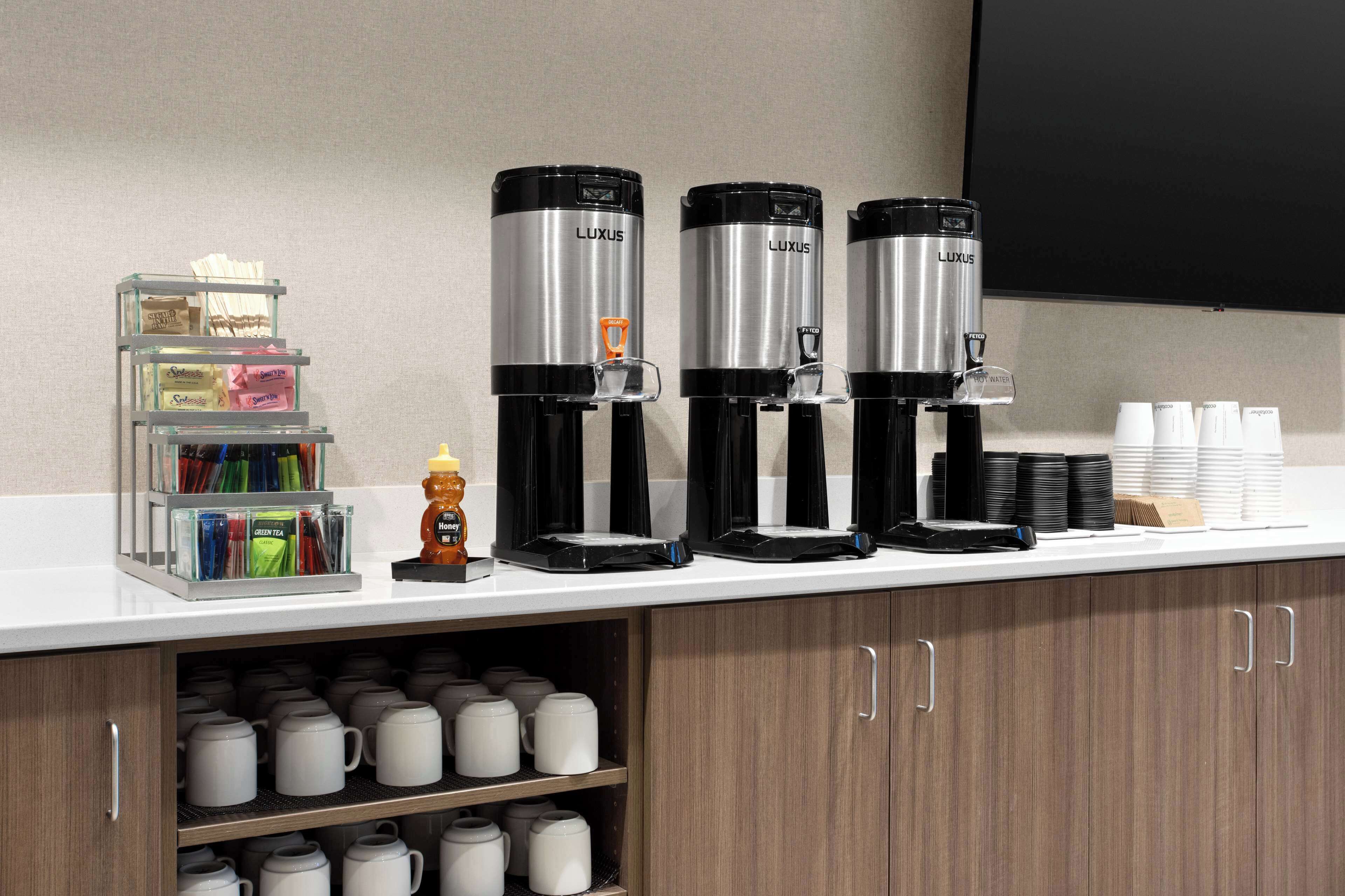 A coffee station with three black and silver coffee makers, a small figurine, and a shelf with mugs and condiments.