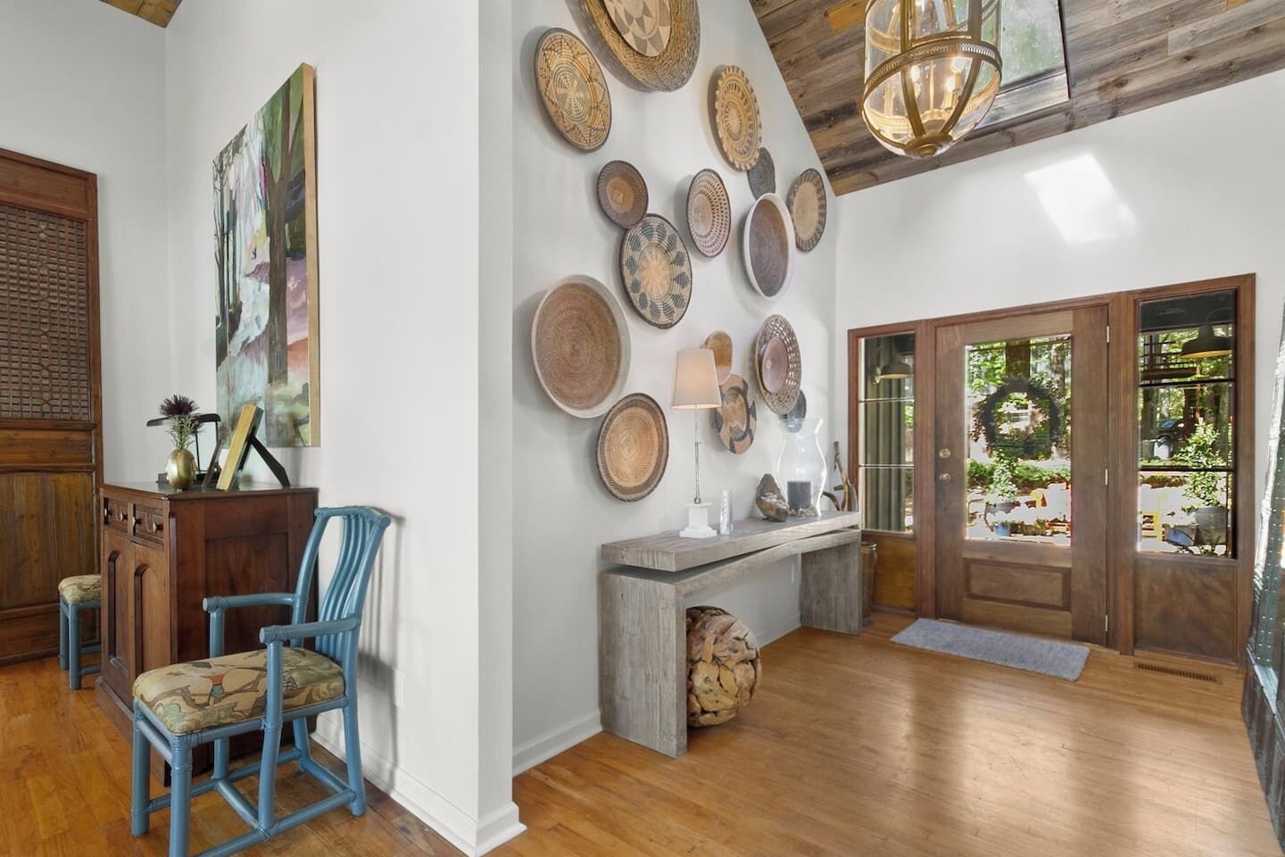 A spacious entryway with a wooden floor, a blue chair, a wooden desk, and a wall adorned with various decorative plates.