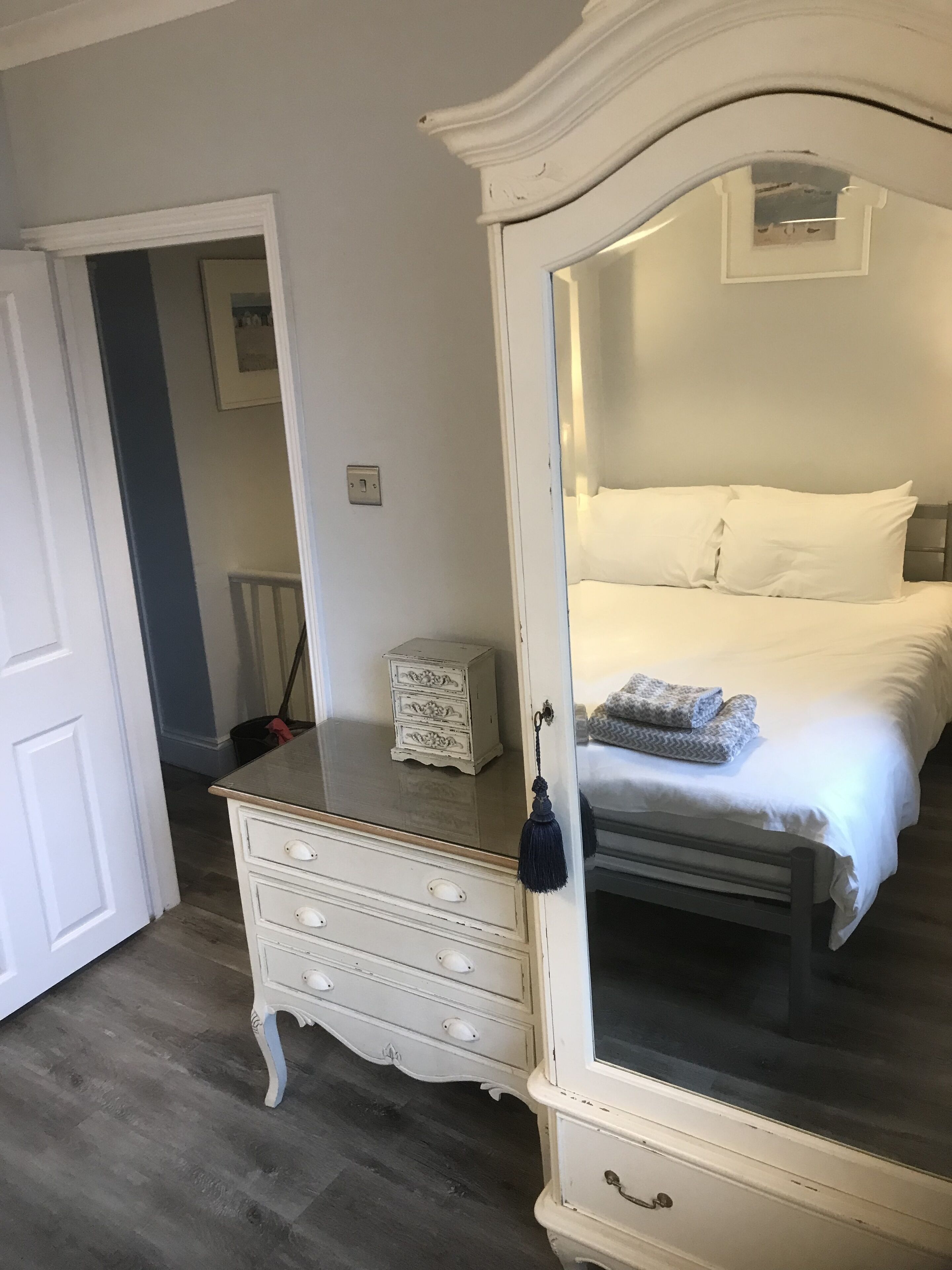 A bedroom with a large mirror, a bed with white linens, a dresser, and a small white chest of drawers.
