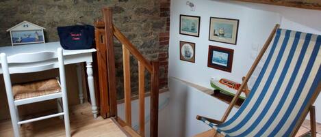 A room with a wooden staircase, a white chair with a striped cushion, a small table with a blue bag, and framed pictures on the wall.