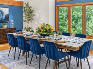 Ein Essbereich mit einem Holztisch, gedeckt für sechs Personen, blauen Stühlen und einem großen Fenster mit Blick auf Grünflächen.