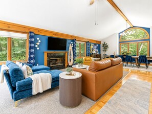 Ein geräumiges Wohnzimmer mit Kamin, blauen und braunen Sofas, einem Fernseher und großen Fenstern mit Blick ins Freie.