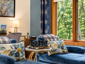 Ein gemütliches Zimmer mit einem blauen Sofa, einem kleinen runden Tisch mit Schachfiguren und einem Fenster mit Blick auf Bäume.