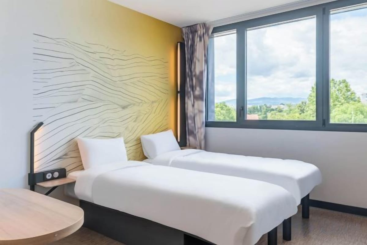 A hotel room with two beds, a desk, a lamp, and a window with a view.