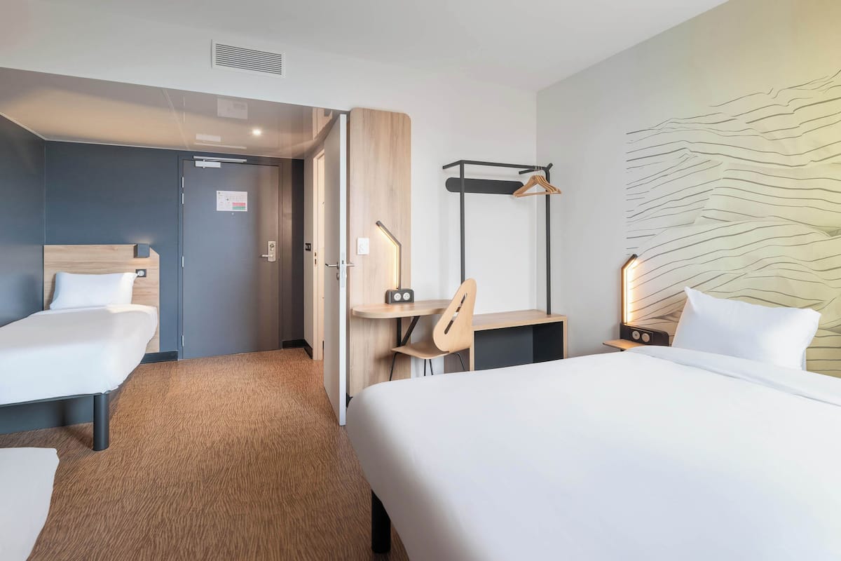 A hotel room with a large bed, two smaller beds, a desk, a chair, and a wall-mounted coat rack.