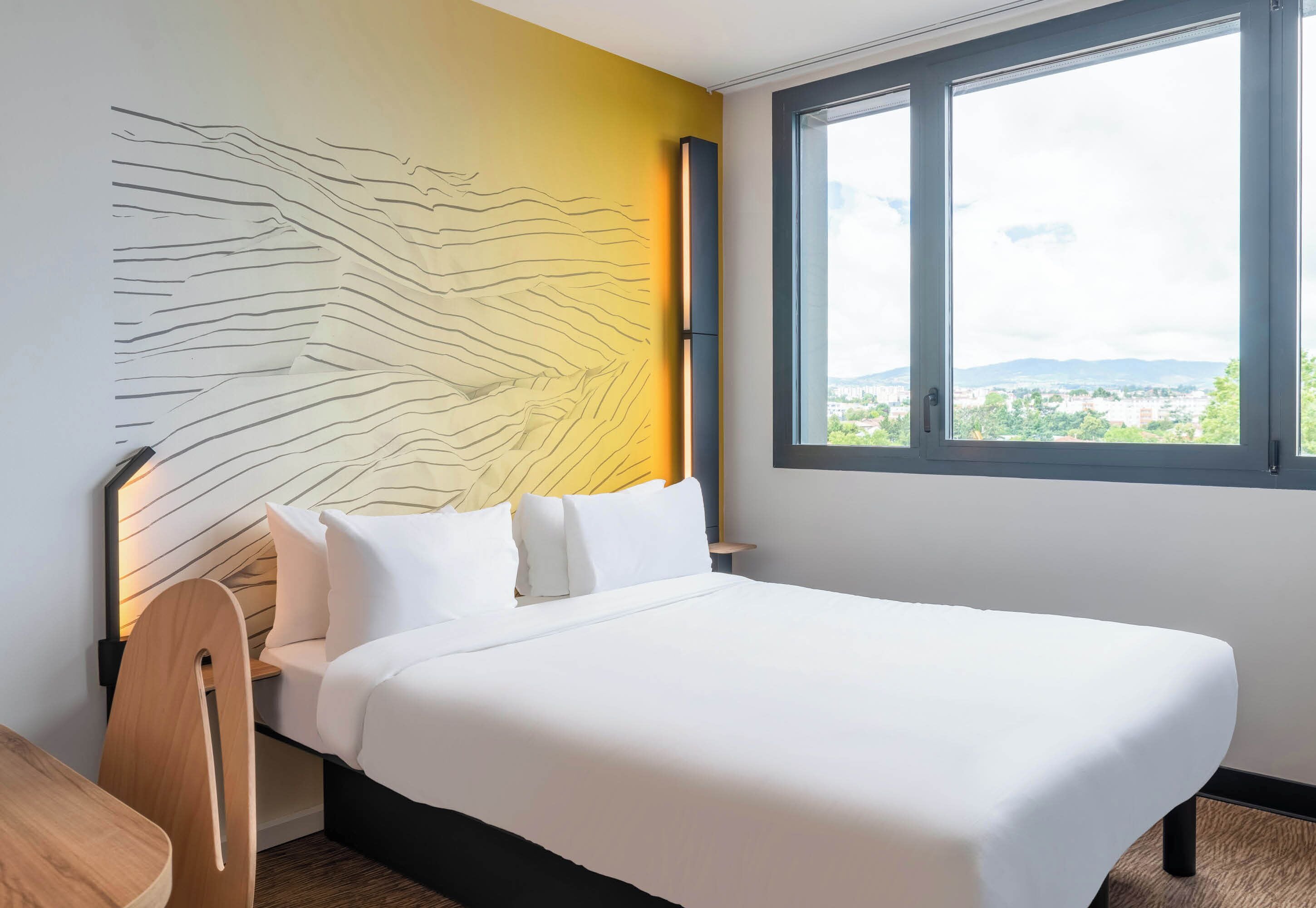 A modern hotel room with a large bed, a headboard with a unique design, a window with a city view, and a wooden chair.