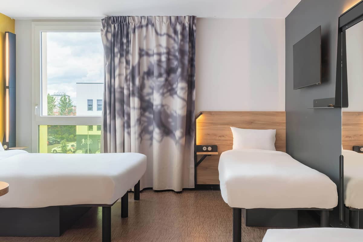 A hotel room with two beds, a large window with curtains, a flat-screen TV mounted on the wall, and a wooden headboard.