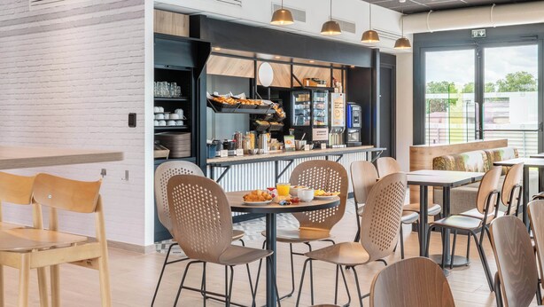 A modern cafe with wooden tables and chairs, a counter with food items, and a view of the outdoors. - B&B HOTEL Villefranche-sur-Saône Sud (Villefranche-sur-Saône)