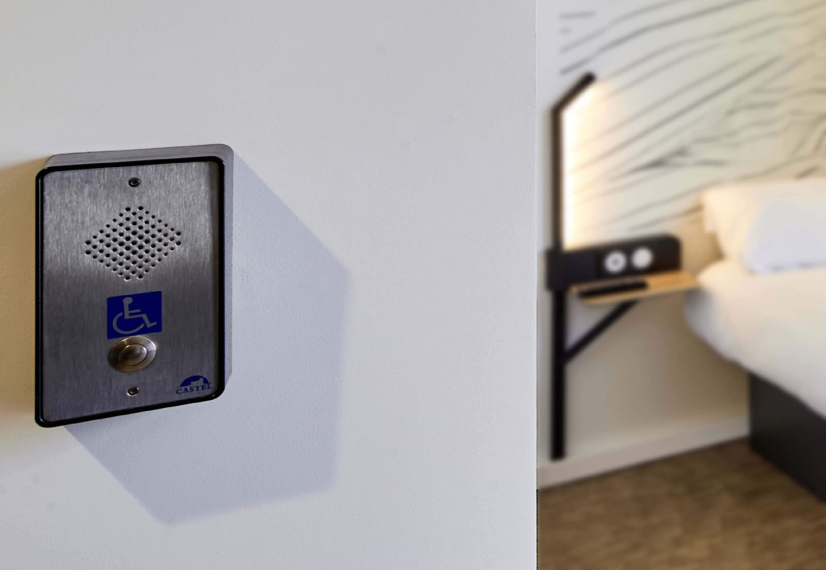 A hotel room with a bed, a nightstand, and a wall-mounted control panel with a wheelchair accessibility symbol.