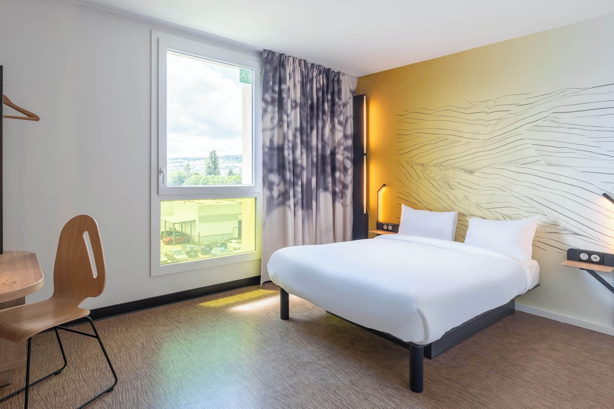 A hotel room with a large bed, a desk with a chair, a window with curtains, and a wall with a textured design.