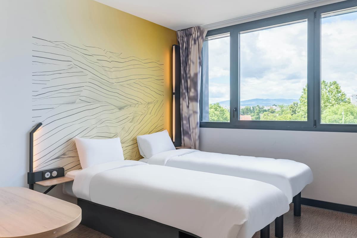 A hotel room with two beds, a wooden table, a headboard with a light pattern, and a window with a view of greenery and mountains.