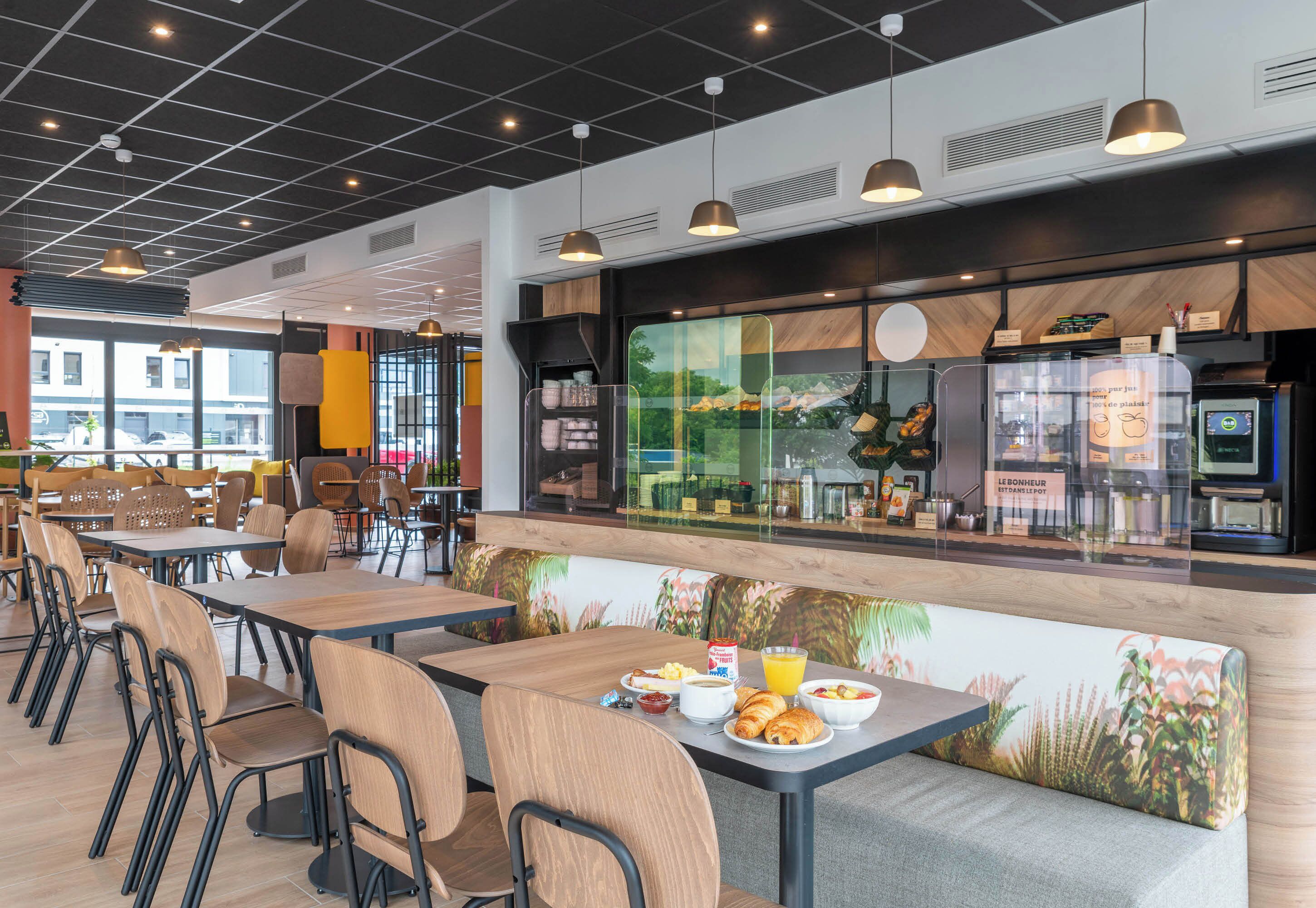 Un café moderno con mesas y sillas de madera, un amplio ventanal con vistas al exterior y una barra con productos alimenticios.