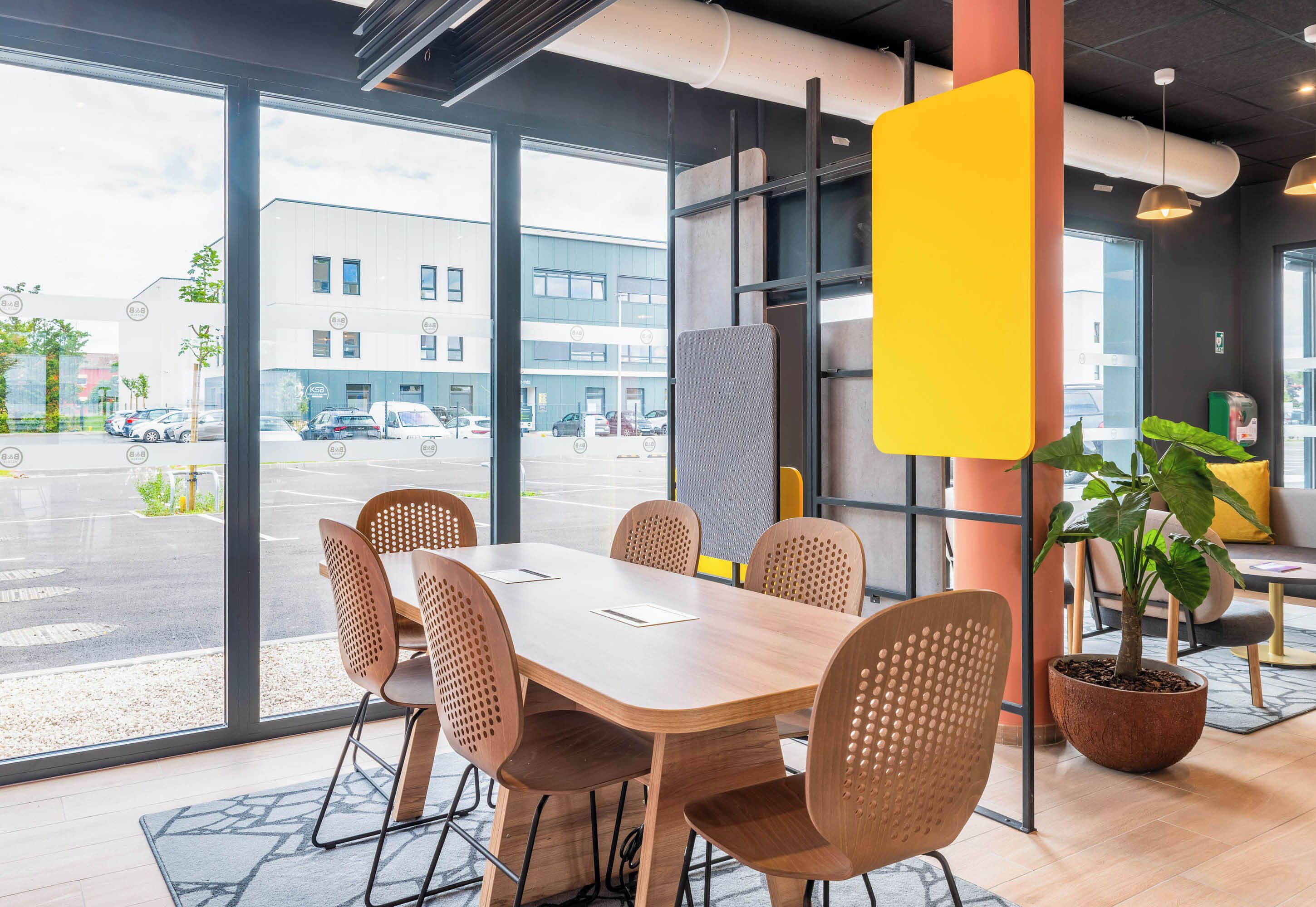 Una sala de conferencias moderna con una mesa grande y seis sillas, ventanas amplias con vista a un estacionamiento y edificios, y una planta en maceta.