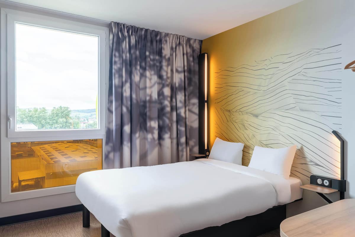A hotel room with a large bed, a bedside table, a nightstand, a window with curtains, and a view of the outdoors.