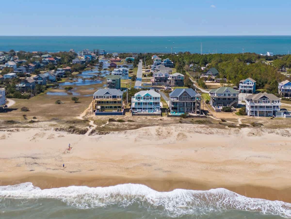 Una zona residencial costera con propiedades frente a la playa, incluyendo casas con amplios ventanales y balcones, una playa arenosa y el océano al fondo.