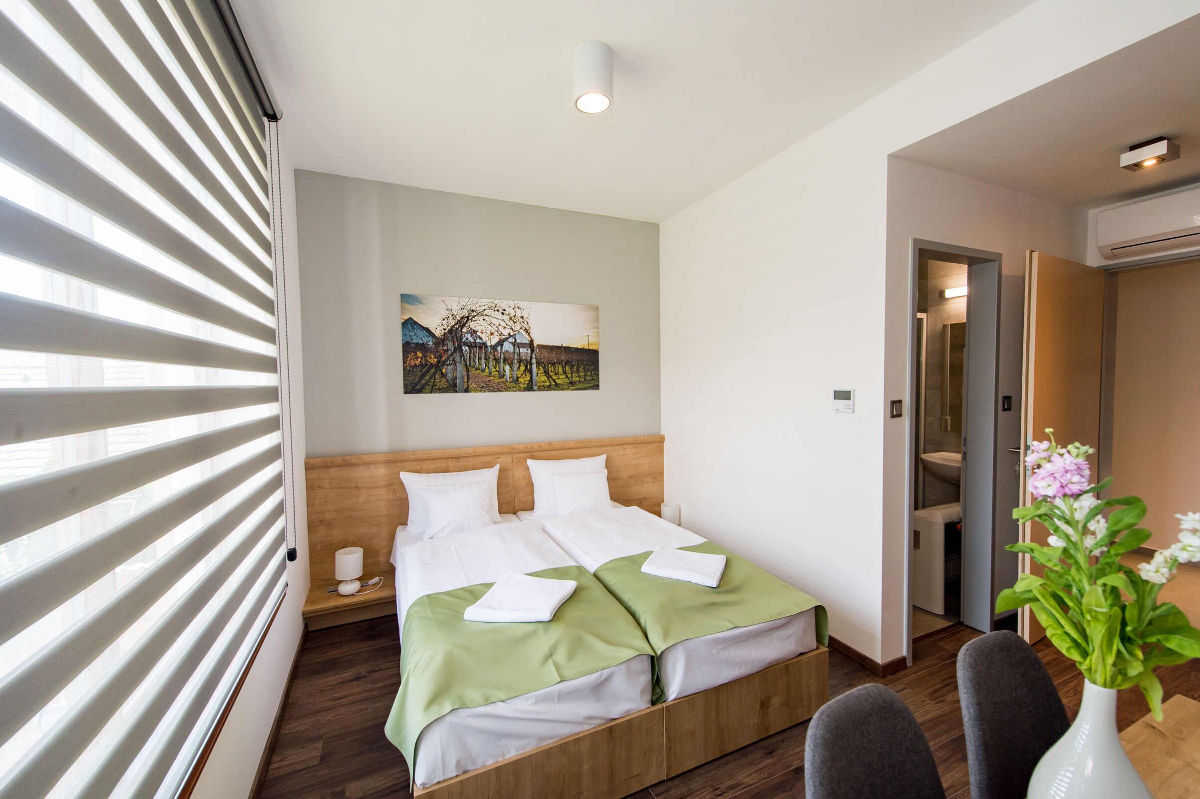 A hotel room with a bed, a painting on the wall, a wooden headboard, a bedside table, a vase with flowers, and a door leading to a bathroom.