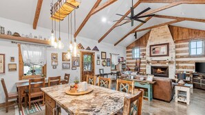 A rustic interior with exposed wooden beams, a stone fireplace, and a dining area with a marble table. - The Outpost Vacation Cabin Fredericksburg TX Pet Friendly Hot Tub Near Enchanted Rock (Fredericksburg)