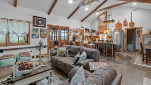 A spacious living room with a large brown sofa, a coffee table with decorative items, and a kitchen area in the background. - The Outpost Vacation Cabin Fredericksburg TX Pet Friendly Hot Tub Near Enchanted Rock (Fredericksburg)