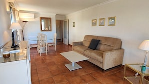 A living room with a beige sofa, a glass coffee table, a white side table, and a dining area with a glass table and chairs. - 50 m² cocoon of Love with a common shared pool (Saint-Raphaël)