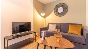Una sala moderna con una televisión de pantalla plana sobre un soporte negro, una mesa de centro redonda de madera con un florero de vidrio, un sofá gris con dos cojines y un espejo colgado en la pared.
