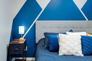 Un dormitorio con diseño geométrico azul y blanco en la pared, cabecera tapizada en gris y mesita de noche con lámpara.