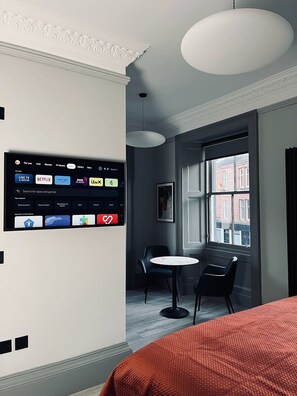 Une chambre d’hôtel moderne équipée d’un téléviseur fixé au mur, permettant d’accéder à diverses applications de diffusion en continu, d’une table ronde, de deux chaises et d’une grande fenêtre donnant sur des immeubles.
