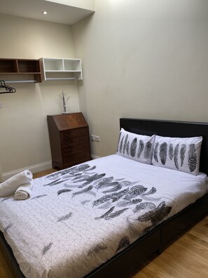 A neatly made bed with a geometric feather pattern bedspread, a dark headboard, and matching pillows. There is a wooden dresser with drawers and a small plant on top. A white towel is rolled on the bed. - Spacious 3-bed Apartment in Central London (London)