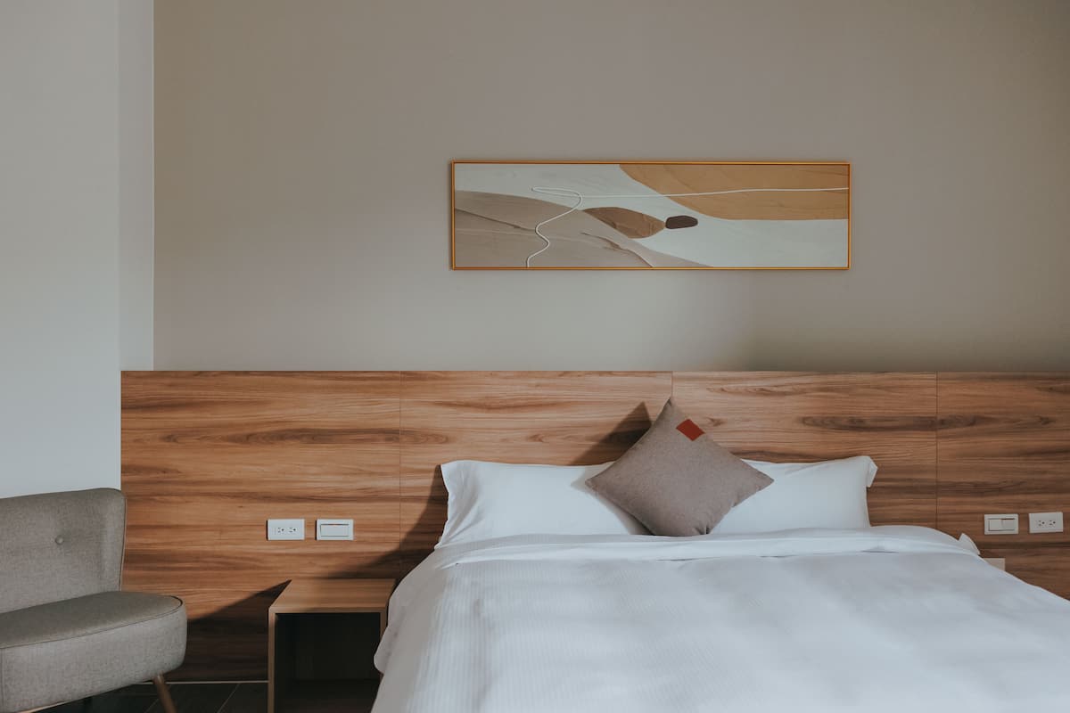 A modern hotel room with a wooden headboard, a bed with white linens, a grey armchair, and a minimalist artwork on the wall.