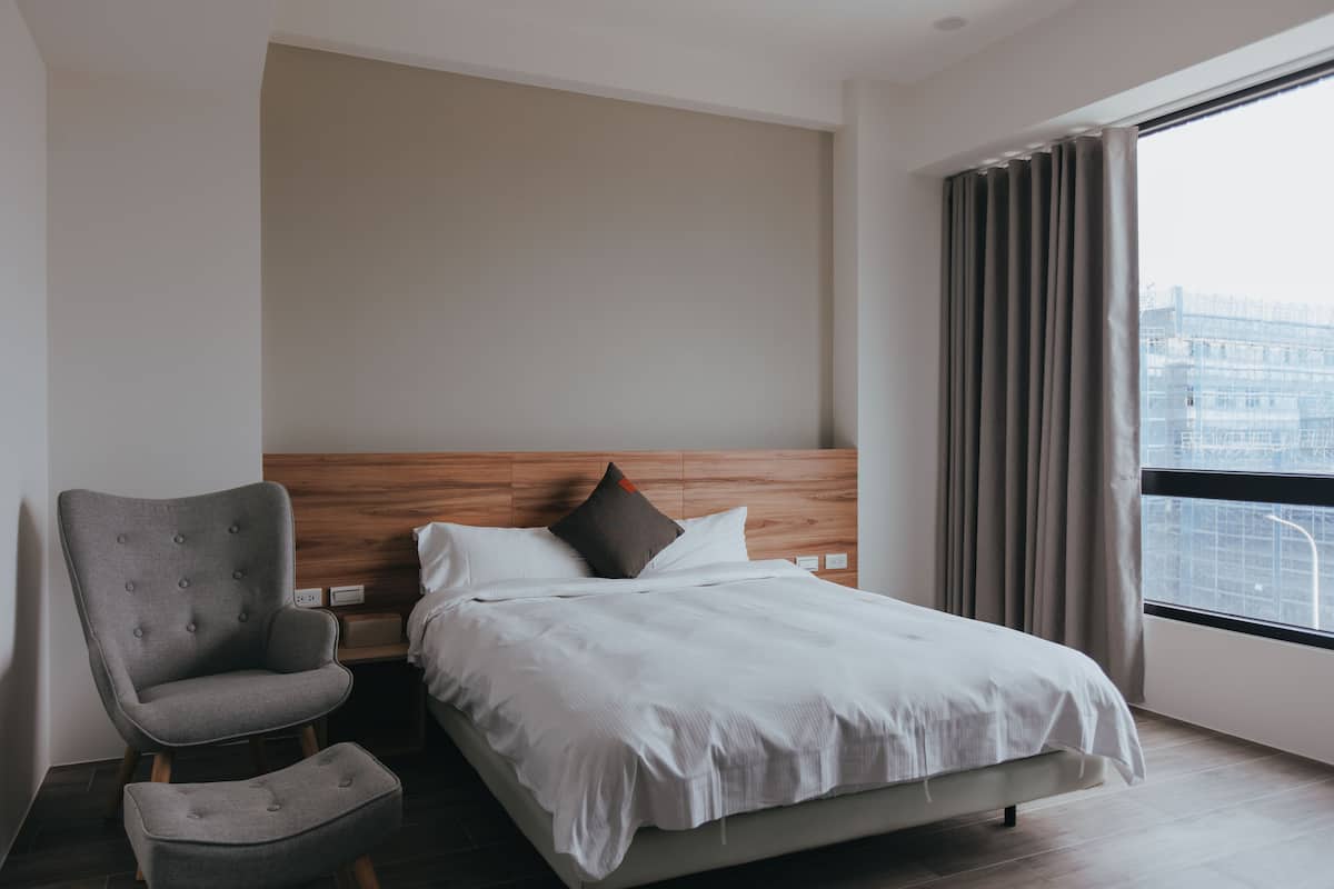 A modern hotel room with a large bed, a grey armchair, a small ottoman, a wooden headboard, and a view of the city through a large window.