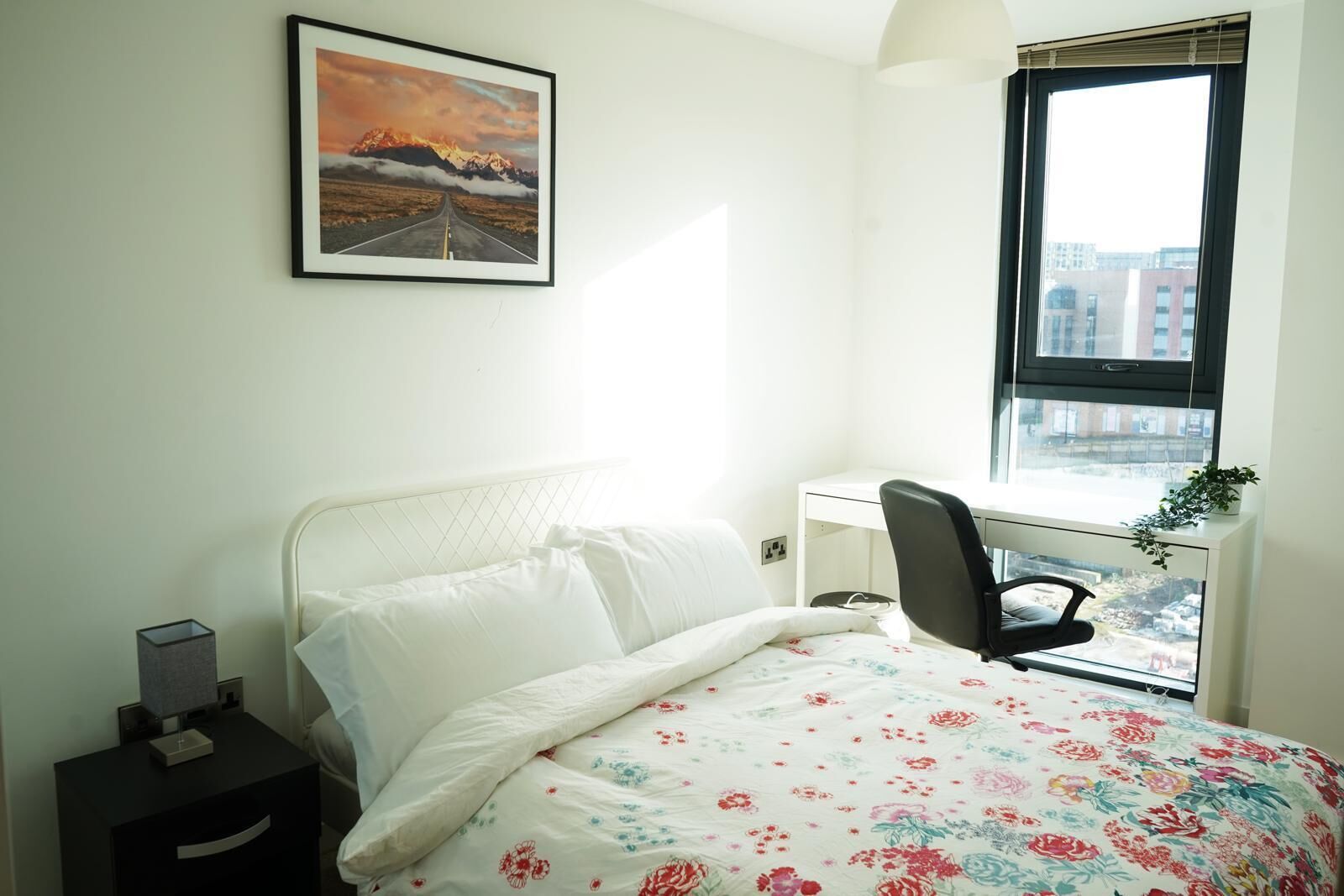 A bedroom with a bed, a desk, and a window with a view of buildings.