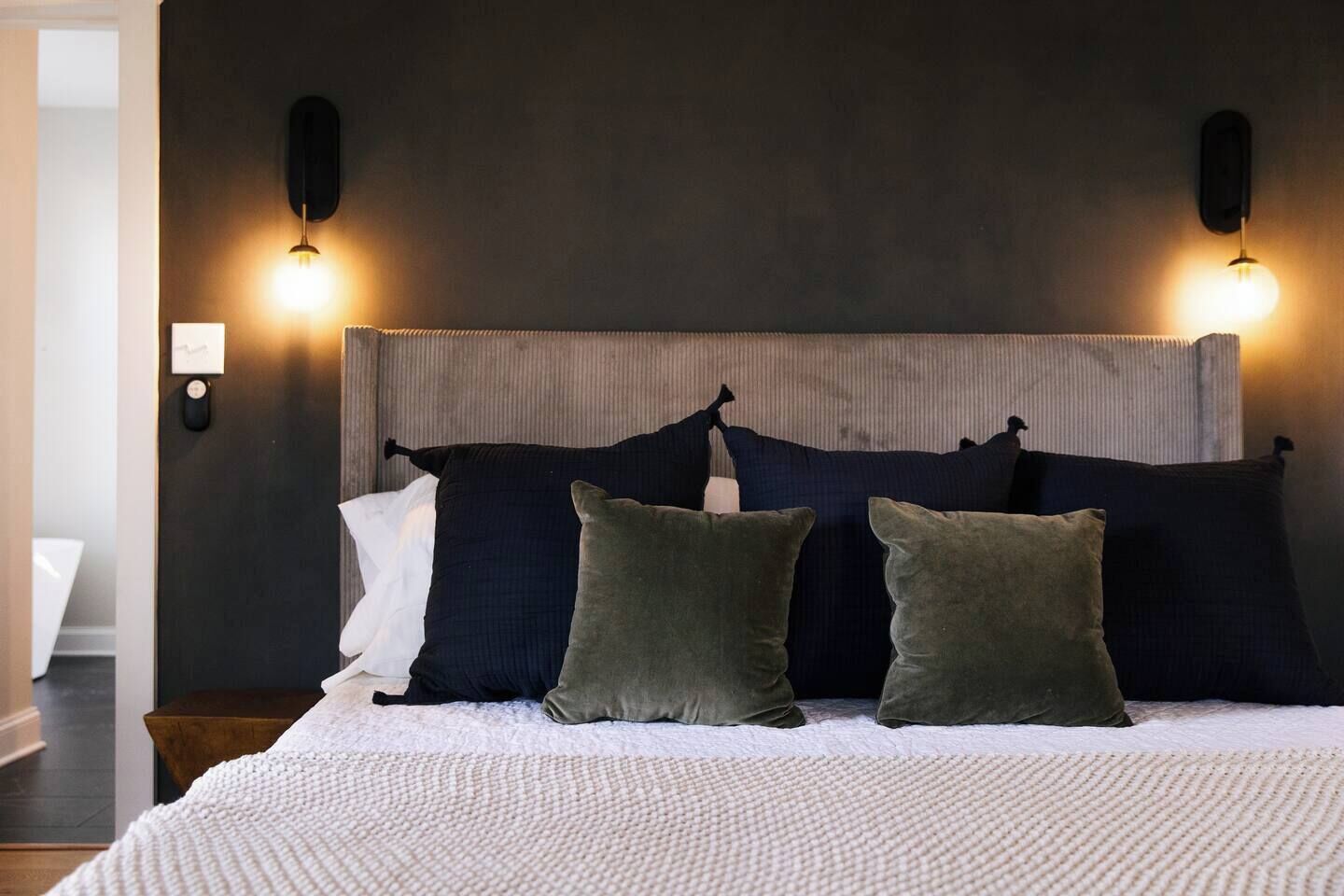 Una cama bien hecha con edredón blanco y varios almohadones de distintos colores.