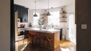 A modern kitchen with a central island, bar stools, pendant lighting, and wooden shelves.
