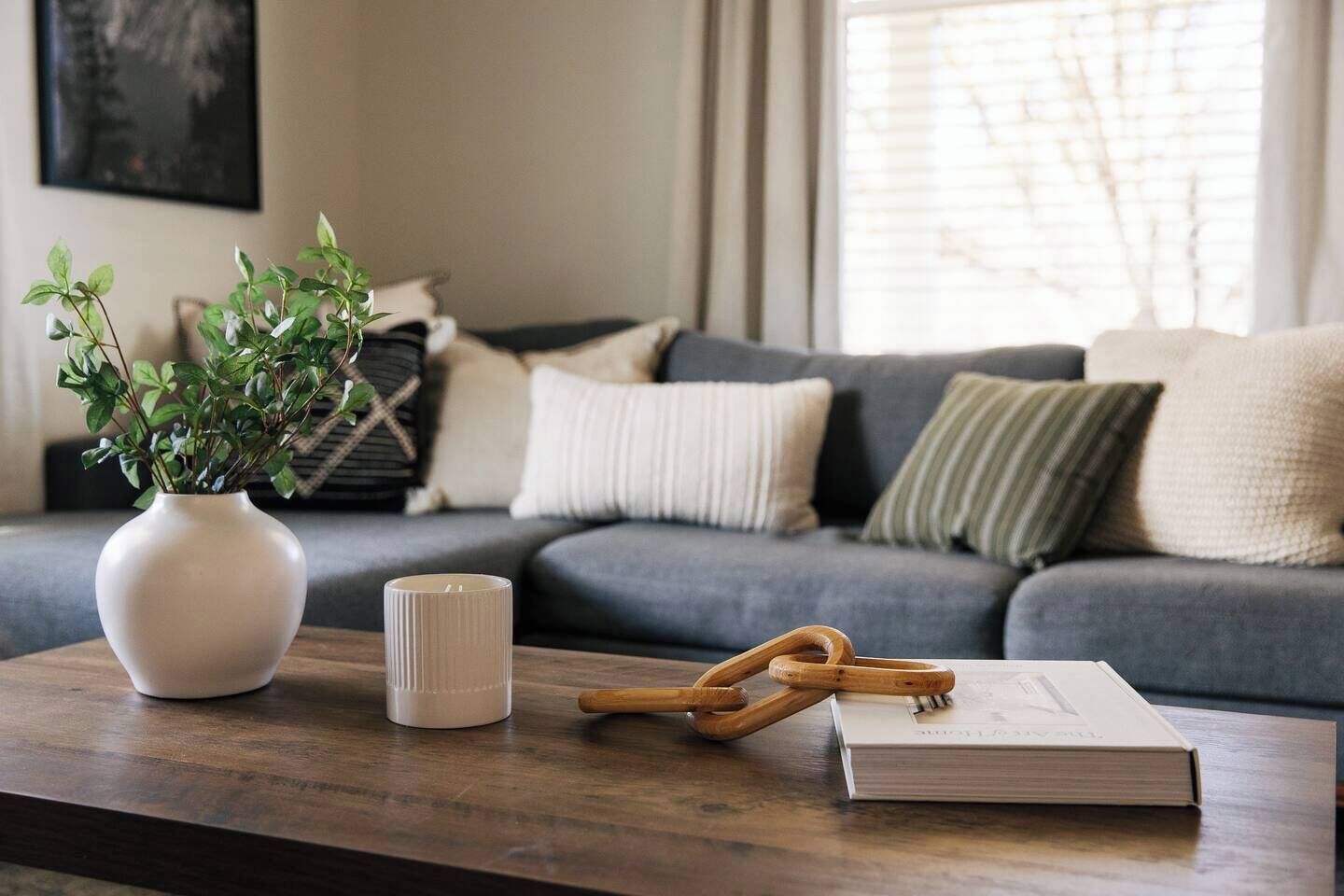 Un salon avec un canapé gris, un vase blanc contenant des plantes vertes, une bougie blanche et un livre posé sur une table en bois.