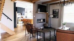 A modern living room with a fireplace, wooden flooring, and a kitchen in the background.