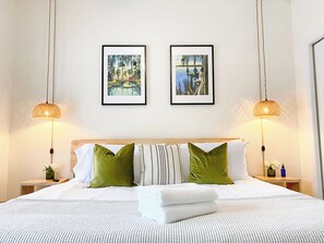 A neatly made bed with white and green pillows, two framed pictures on the wall, and two hanging lamps. - The Mangrove Suite. Newly Built, Upscale, Central (Gainesville)