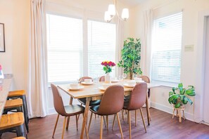 Un coin repas avec une table en bois dressée pour quatre personnes, entourée de chaises, et agrémenté d’une plante et d’un vase de fleurs posés sur la table.