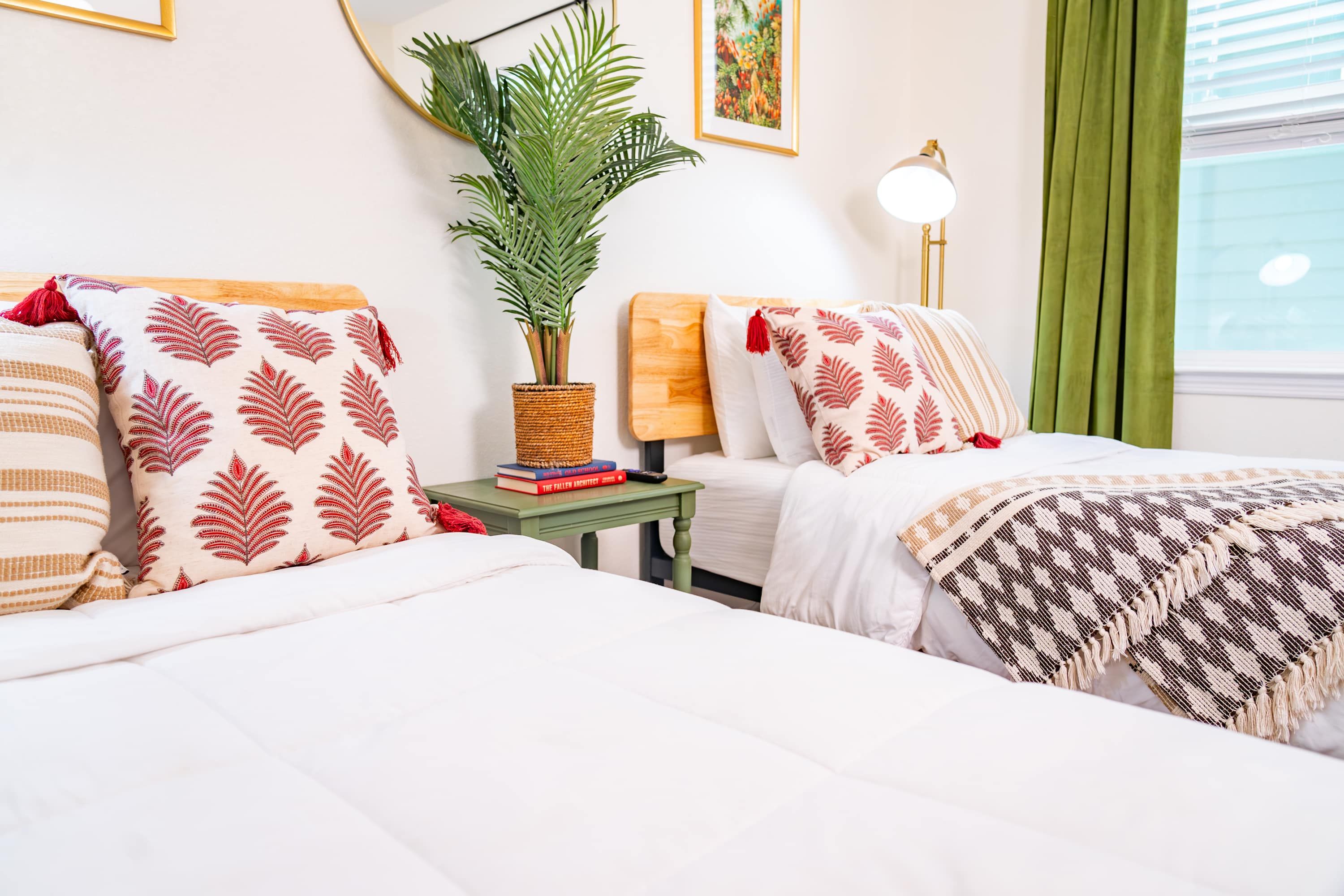A hotel room with two beds, a small table with a plant and books, a lamp, and a window with curtains.