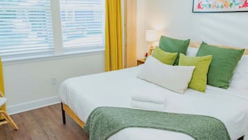 A neatly made bed with white and green bedding, a bedside lamp, and a window with blinds.