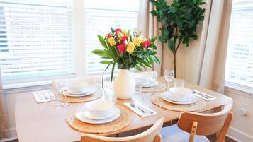 Une table à manger dressée pour deux personnes, avec de l’assiette blanche, des couverts et des verres, un vase de fleurs et une plante en pot.