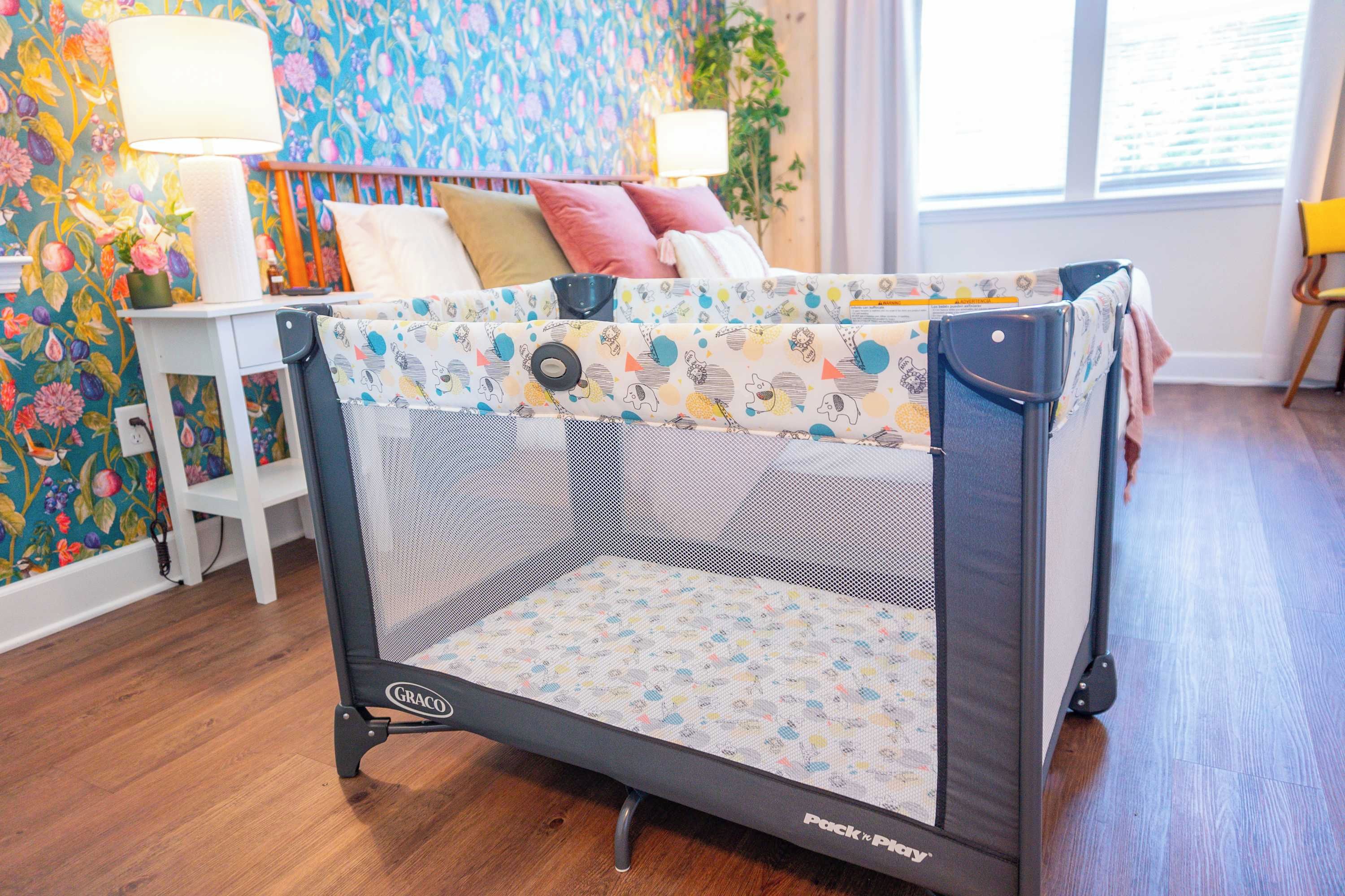 Un parc pour bébé doté d’une partie supérieure en tissu à mailles et d’une partie inférieure à motifs, placé dans une pièce dont le papier peint est à fleurs, le plancher en bois et la fenêtre est équipée de stores.