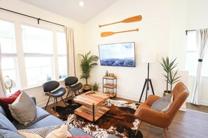 A modern living room with a sofa, chairs, a coffee table, and a cowhide rug. There are windows, a TV, and a wall-mounted shelf with decorative items.
