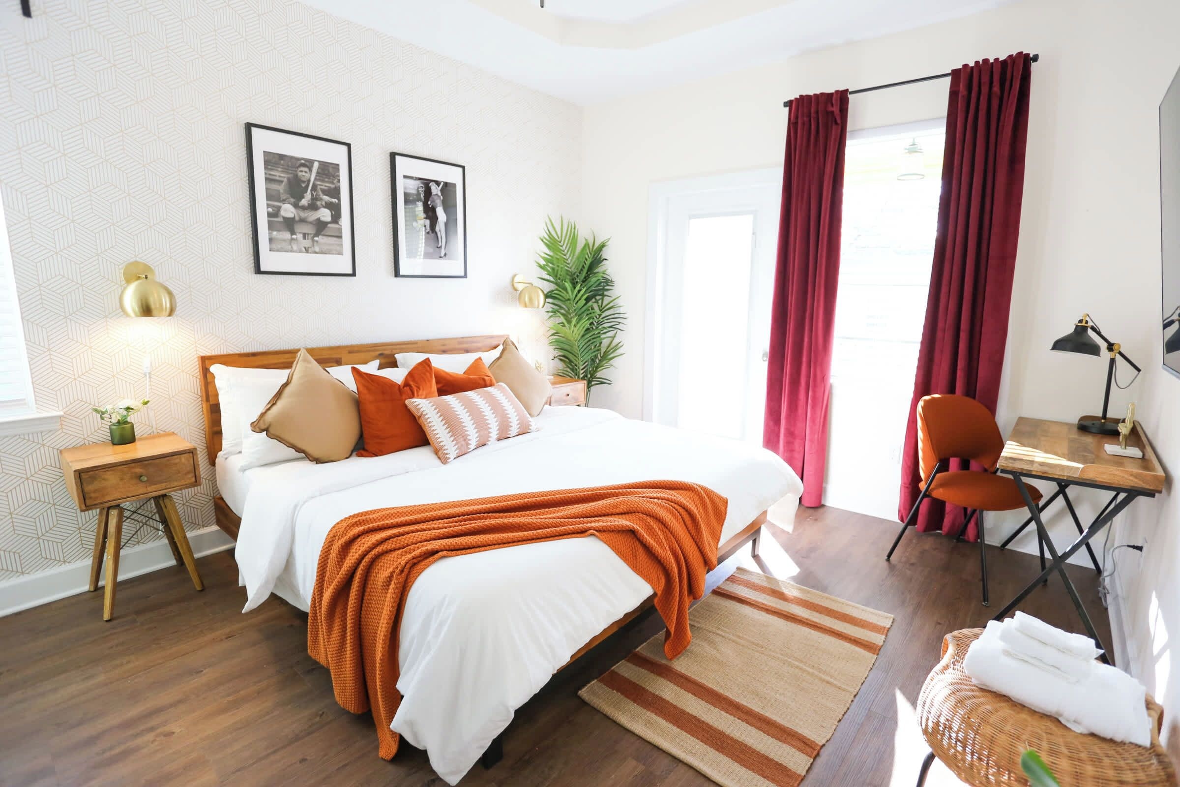 A neatly arranged bedroom with a large bed, a desk, and a chair. There are two framed pictures on the wall, a potted plant, and a window with curtains.