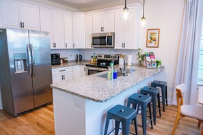 A modern kitchen with white cabinets, stainless steel appliances, a granite countertop, and bar stools. - The Royal Crane Suite, New Build, Upscale, Central (Gainesville)