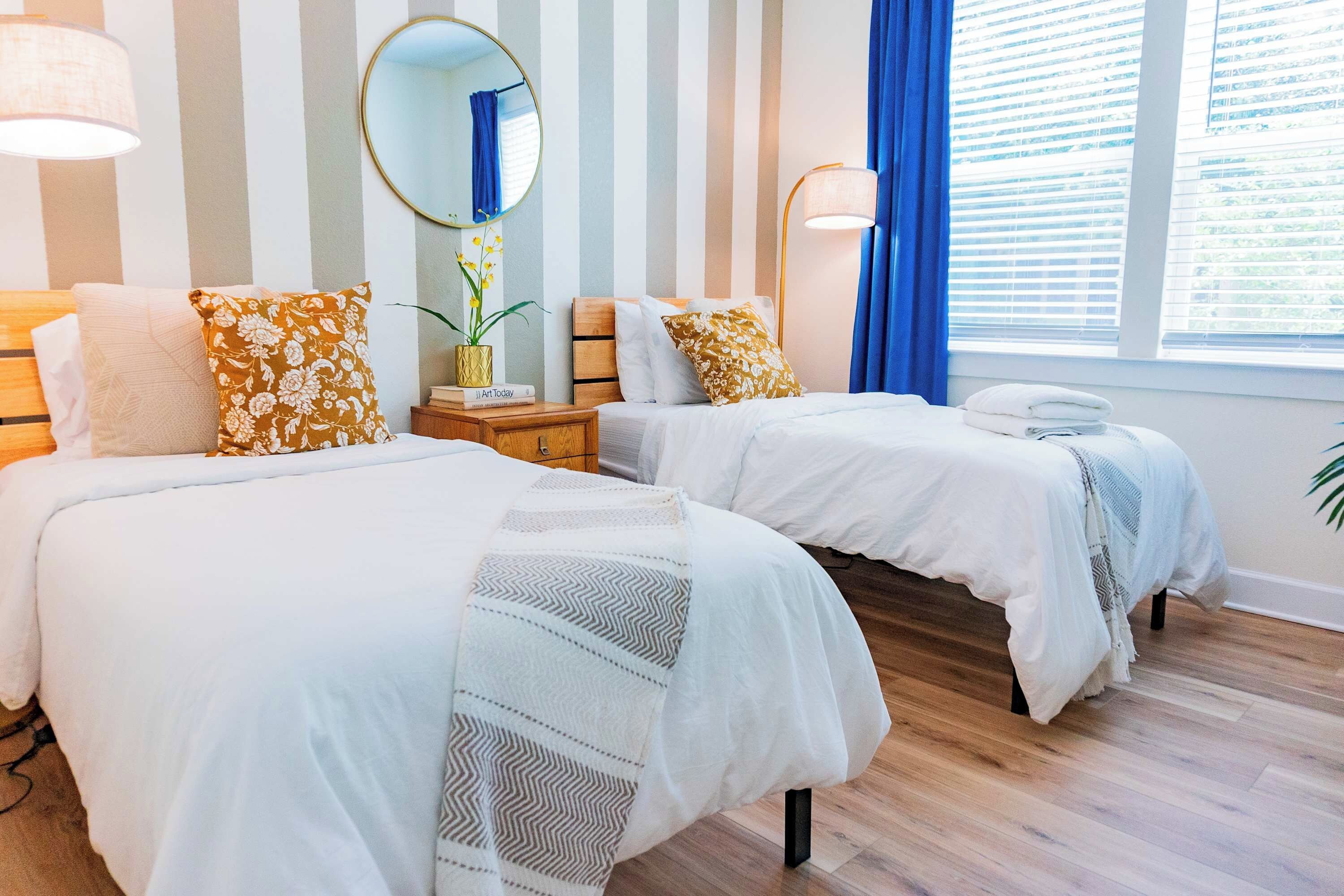Et hotelværelse med to senge, stribet tapet, et rundt spejl, et natbord med lampe og et vindue med gardiner.