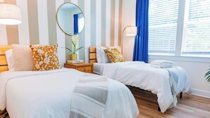 Et hotelværelse med to senge, stribet tapet, et rundt spejl, et natbord med lampe og et vindue med gardiner.