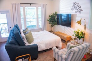 A bedroom with a bed, a blue armchair, a striped armchair, a TV, a plant, and a window with curtains. - The Lotus Suite. Newly Built, Upscale, Central (Gainesville)