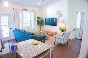 Uma sala de estar moderna com um sofá azul, uma poltrona listrada, uma TV de tela plana, um espaço de jantar com bancada de granito e uma planta em vaso.