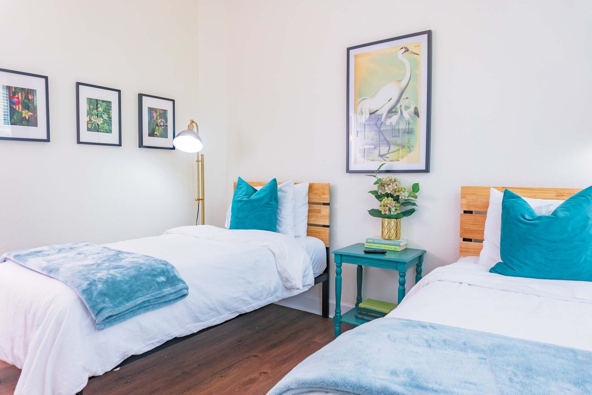 Habitación de hotel con dos camas, una mesita de noche con lámpara, una obra de arte enmarcada en la pared y una pequeña mesa con un jarrón de flores.